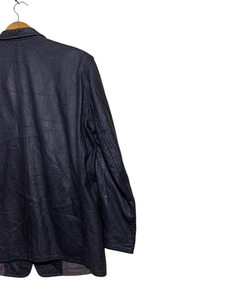 May include: A black leather jacket with a textured finish. The jacket has a classic design with a notched collar and a button closure. The sleeves are long, and the jacket appears to be in good condition. The jacket is hanging on a wooden hanger.