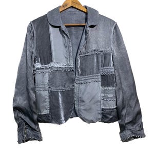 May include: A grey patchwork jacket with a collar and long sleeves. The jacket is made of various grey fabrics, including velvet and satin, and is decorated with lace and small pom-poms. The jacket is hanging on a wooden hanger.