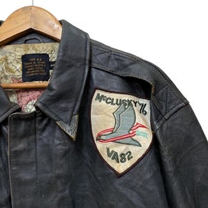 Może przedstawiać: Vintage brązowa sk&oacute;rzana kurtka typu A-2 z naszywką "McClusky 76 VA82". Naszywka przedstawia stylizowanego ptaka i tekst. Kurtka ma kołnierz i podszewkę z wzorem mapy.