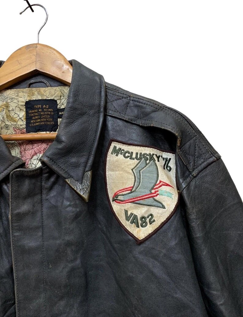 Może przedstawiać: Vintage brązowa sk&oacute;rzana kurtka typu A-2 z naszywką "McClusky 76 VA82". Naszywka przedstawia stylizowanego ptaka i tekst. Kurtka ma kołnierz i podszewkę z wzorem mapy.