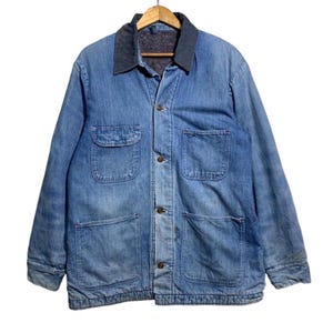 May include: A light blue denim chore coat featuring a dark grey corduroy collar, long sleeves, and four front patch pockets. The jacket is secured with four brown buttons down the center front and is displayed on a wooden hanger against a white background.