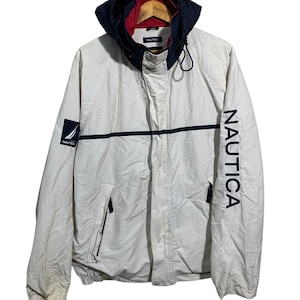 May include: Off-white Nautica jacket with a navy blue and red lined hood. The jacket features a navy blue horizontal stripe across the chest and the word "NAUTICA" printed vertically on the right sleeve. A small Nautica logo patch is on the left chest.