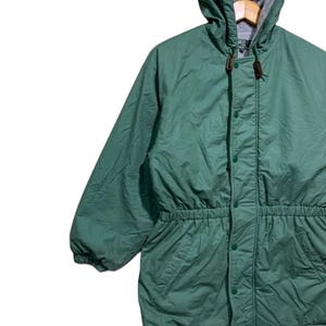 Pu&ograve; includere: Giacca con cappuccio verde con vita regolabile e chiusura con bottoni. La giacca ha un cappuccio foderato in pile grigio e un toggle in pelle marrone. La giacca &egrave; realizzata in un materiale leggero.