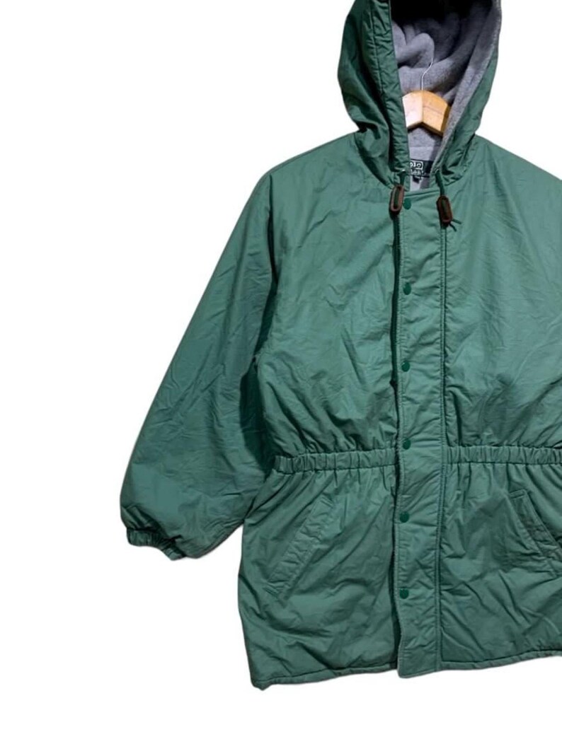 Pu&ograve; includere: Giacca con cappuccio verde con vita regolabile e chiusura con bottoni. La giacca ha un cappuccio foderato in pile grigio e un toggle in pelle marrone. La giacca &egrave; realizzata in un materiale leggero.