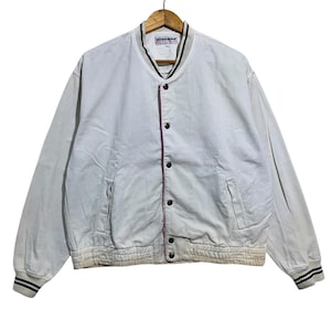 May include: White vintage bomber jacket with black buttons, a black and white striped cuff, and a collar with green and black trim. The jacket has two front pockets and a purple accent along the button line. The brand name "MOSHKIMA" is visible on the collar.