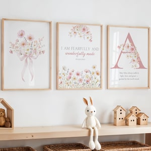 Puede incluir: Tres impresiones artísticas florales enmarcadas con marcos de madera cuelgan de una pared blanca. Las impresiones presentan flores de acuarela y citas inspiradoras. Una estantería de madera debajo muestra un oso de peluche en un marco con forma de casa, un conejito de peluche y casas decorativas de madera.