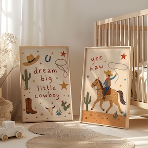 May include: Two framed art prints with a cowboy theme. One features a cowboy hat, boot, cactus, and the text "dream big little cowboy." The other shows a cowboy riding a horse with the text "yee haw." Both have a light wood frame.