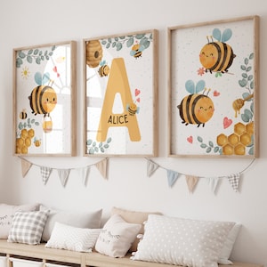 May include: Three framed prints with a watercolor illustration of bees and honeycombs. The prints feature the letter A, the name Alice, and a bee with a happy face. The prints are on a white wall with a garland of flags.