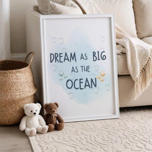 May include: A framed print with the text "DREAM AS BIG AS THE OCEAN" in navy blue, with a watercolour-style blue background and bubble and fish illustrations. Two teddy bears and a woven basket are in the foreground.