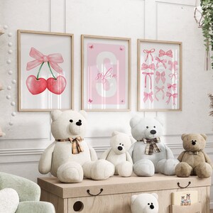 May include: Three framed prints with pink and white watercolor illustrations of cherries, a bow, and the name "sofia" in a pink and white font. Three teddy bears are sitting on a wooden shelf in front of the prints.