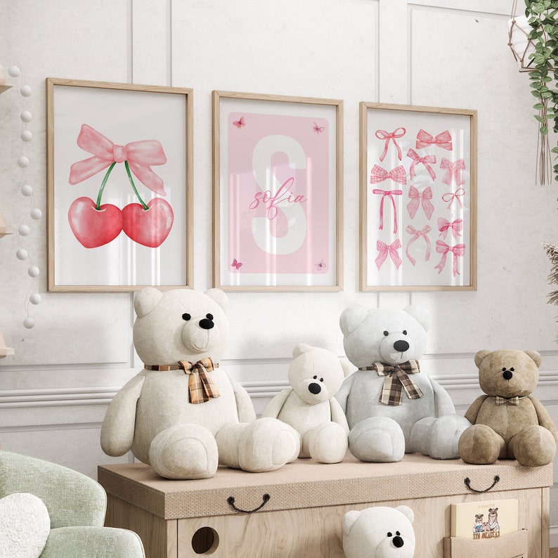 May include: Three framed prints with pink and white watercolor illustrations of cherries, a bow, and the name "sofia" in a pink and white font. Three teddy bears are sitting on a wooden shelf in front of the prints.