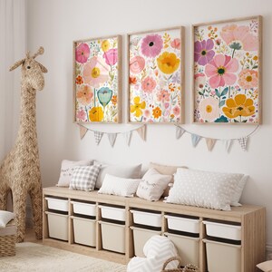 May include: Three framed floral art prints with colorful watercolor flowers in pink, yellow, and orange hues. The prints are mounted on a white wall above a wooden storage bench with white bins. A woven giraffe and pillows complete the room.