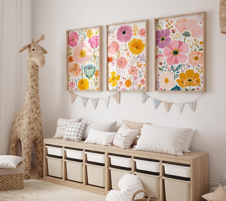 May include: Three framed floral art prints with colorful watercolor flowers in pink, yellow, and orange hues. The prints are mounted on a white wall above a wooden storage bench with white bins. A woven giraffe and pillows complete the room.