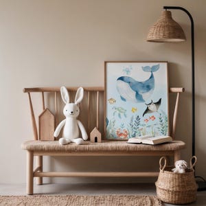 May include: A wooden bench with a woven seat displays a framed watercolour print of a whale and marine life. A white stuffed bunny, wooden houses, and a woven basket with a small stuffed dog are also present.