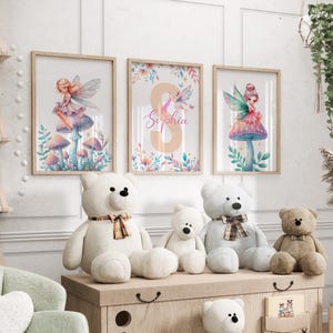 Peut inclure: Trois impressions encadr&eacute;es avec des illustrations &agrave; l'aquarelle de f&eacute;es assises sur des champignons. Les impressions ont une palette de couleurs rose p&acirc;le, bleu et vert. L'impression du milieu pr&eacute;sente le nom "Sophia" en lettres roses. Les impressions sont affich&eacute;es sur un mur au-dessus d'une &eacute;tag&egrave;re en bois avec des animaux en peluche.