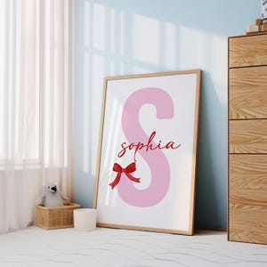 May include: A pink letter S with the name "sophia" written in cursive below it. A red bow is at the bottom of the letter. The print is framed in a light wood frame.