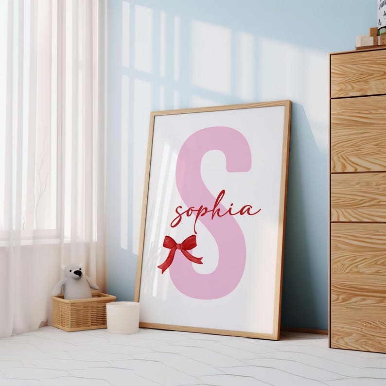 May include: A pink letter S with the name "sophia" written in cursive below it. A red bow is at the bottom of the letter. The print is framed in a light wood frame.