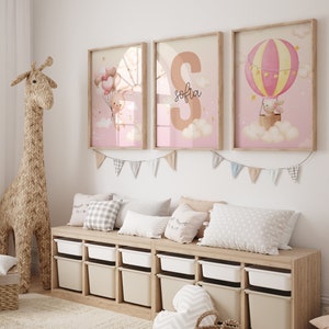 Pu&ograve; includere: Tre stampe incorniciate con uno schema di colori rosa e bianco. Le stampe raffigurano un coniglio che tiene dei palloncini, una grande lettera "S" con il nome "Sofia" scritto sotto, e un coniglio che cavalca una mongolfiera. Le stampe sono appese a un muro bianco sopra uno scaffale di legno con contenitori di stoccaggio.