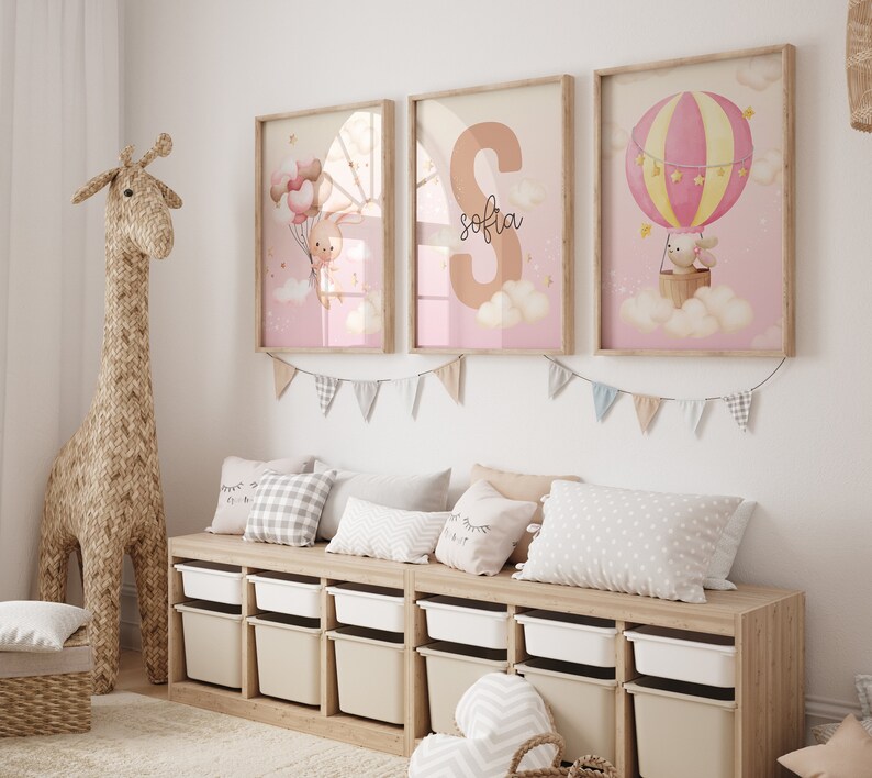 Pu&ograve; includere: Tre stampe incorniciate con uno schema di colori rosa e bianco. Le stampe raffigurano un coniglio che tiene dei palloncini, una grande lettera "S" con il nome "Sofia" scritto sotto, e un coniglio che cavalca una mongolfiera. Le stampe sono appese a un muro bianco sopra uno scaffale di legno con contenitori di stoccaggio.