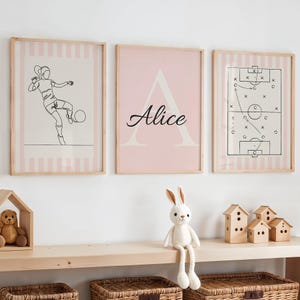 Op de afbeelding: Drie ingelijste prints met een voetbalthema. Eén toont een lijntekening van een voetballer, een andere heeft de naam "Alice" met een grote letter "A", en de derde toont een voetbalspeldiagram. Alle zijn in lichtroze en wit.