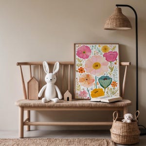 May include: A framed floral print with pink, yellow, and orange flowers sits on a wooden bench with a woven seat. A white bunny plush and two wooden houses are on the bench. A woven basket and a floor lamp are also in the scene.