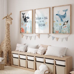 May include: Three framed watercolour prints with ocean themes hang above a storage bench. The prints feature whales, an orca, and sea creatures. The middle print has the text "Dream as big as the ocean". A woven giraffe and pillows complete the room.