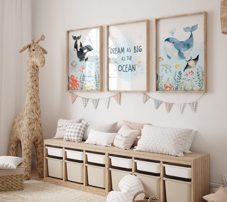 May include: Three framed watercolour prints with ocean themes hang above a storage bench. The prints feature whales, an orca, and sea creatures. The middle print has the text "Dream as big as the ocean". A woven giraffe and pillows complete the room.
