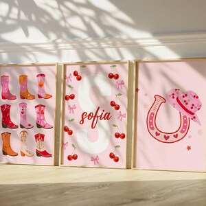 K&ouml;nnte beinhalten: Drei gerahmte rosa Drucke mit Western-Motiv. Ein Druck zeigt verschiedene Cowboy-Stiefel, ein anderer den Namen "Sofia" mit Kirschen, und der letzte ein Hufeisen und einen Cowboyhut. Die Drucke haben einen hellrosa Hintergrund.