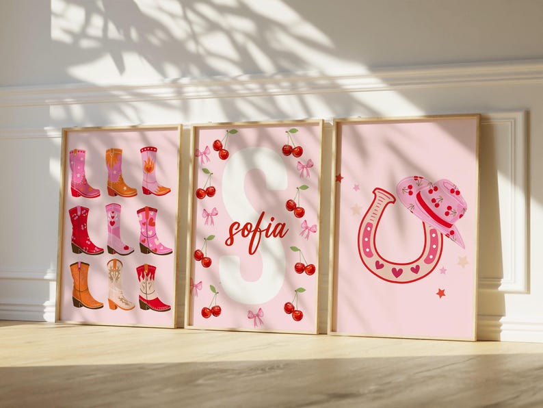 K&ouml;nnte beinhalten: Drei gerahmte rosa Drucke mit Western-Motiv. Ein Druck zeigt verschiedene Cowboy-Stiefel, ein anderer den Namen "Sofia" mit Kirschen, und der letzte ein Hufeisen und einen Cowboyhut. Die Drucke haben einen hellrosa Hintergrund.