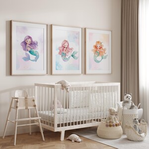 May include: Three watercolor prints of mermaids with different colored hair and tails. The prints are framed in light wood and hung on a white wall above a white crib.
