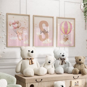 Pu&ograve; includere: Tre stampe incorniciate con sfondo rosa e un tema fantasioso. Le stampe mostrano un coniglio che tiene dei palloncini, la lettera 'S' con il nome 'sofia' scritto sotto, e una mongolfiera con un coniglio dentro. Le stampe sono esposte su una parete bianca sopra uno scaffale di legno con quattro orsacchiotti.