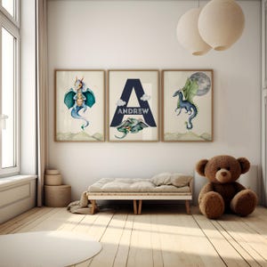 May include: Three framed prints with watercolor illustrations of dragons. The middle print features the letter "A" in a navy blue triangle with the name "ANDREW" written below it. The prints are hanging on a white wall in a child's bedroom.