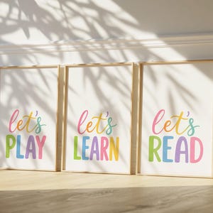 May include: Three framed art prints with the words "Let's PLAY", "Let's LEARN", and "Let's READ" in colorful, playful lettering. The prints are in wooden frames and lean against a white wall, with sunlight casting shadows.