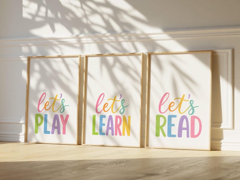 May include: Three framed art prints with the words "Let's PLAY", "Let's LEARN", and "Let's READ" in colorful, playful lettering. The prints are in wooden frames and lean against a white wall, with sunlight casting shadows.