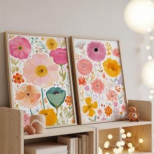May include: Two framed floral art prints. The prints feature watercolor flowers in shades of pink, orange, yellow, and teal against a white background. The frames are made of light-colored wood. The artwork is displayed on a wooden shelf.