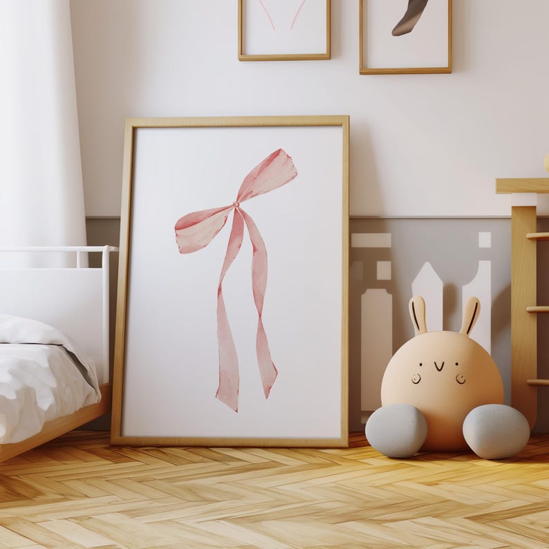 May include: A framed print of a pink watercolor bow on a white background. The frame is made of wood and has a light brown finish.