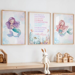 May include: Three framed mermaid art prints. The prints feature watercolor illustrations of mermaids with purple and pink hair, and teal tails. One print has text that reads "I'M UNIQUE, I'M STRONG, I'M SMART, I'M BRAVE, I'M SUPPORTED, I'M HONEST, I'M KIND, I'M BEAUTIFUL, I'M ENOUGH, I'M LOVED, I AM Alice."