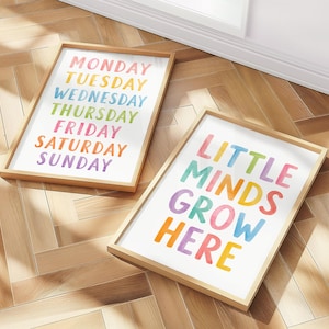 May include: Two framed art prints with wooden frames. One print displays the days of the week in rainbow colors. The other print says "LITTLE MINDS GROW HERE" in colorful letters. The prints are on a light wood floor.