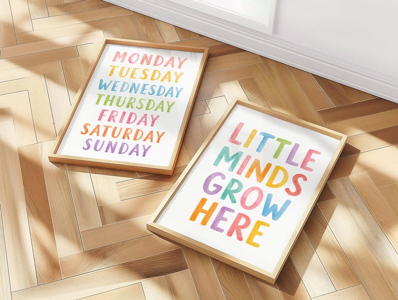 May include: Two framed art prints with wooden frames. One print displays the days of the week in rainbow colors. The other print says "LITTLE MINDS GROW HERE" in colorful letters. The prints are on a light wood floor.