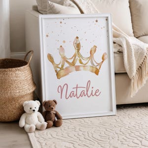 May include: A framed print featuring a watercolor illustration of a gold and pink crown with the name "Natalie" in pink script. The artwork is set against a white background with gold star accents. Two teddy bears and a woven basket are in the foreground.
