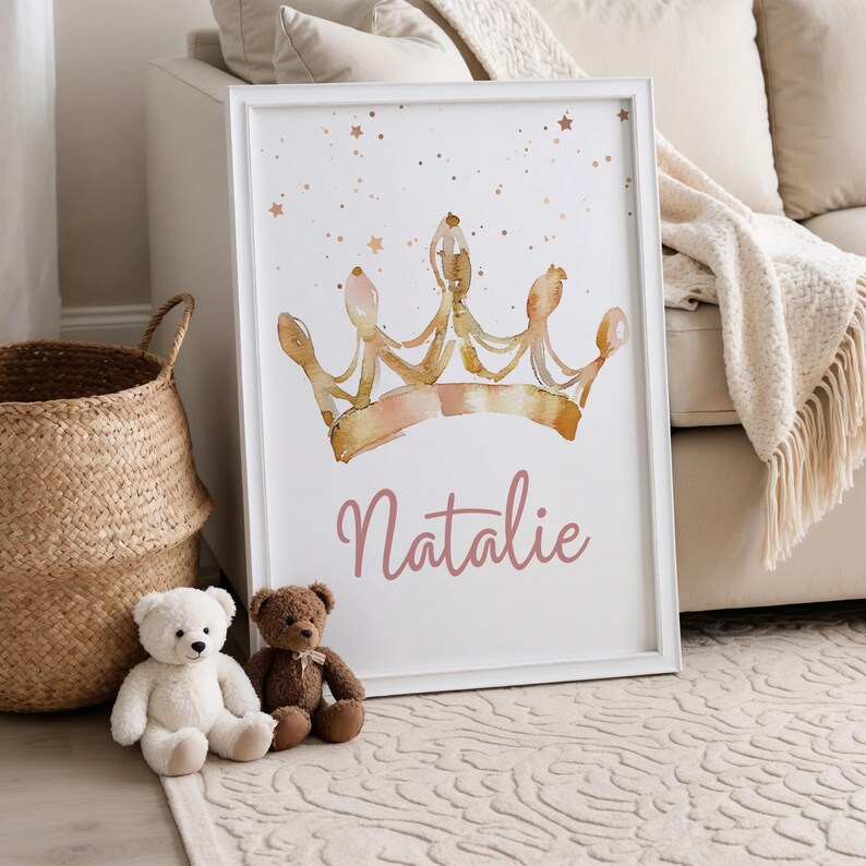 May include: A framed print featuring a watercolor illustration of a gold and pink crown with the name "Natalie" in pink script. The artwork is set against a white background with gold star accents. Two teddy bears and a woven basket are in the foreground.