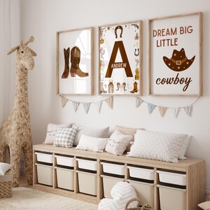 May include: Three framed prints with a western theme. The first print features a pair of brown cowboy boots. The second print features the letter A with the name Andrew below it. The third print features a cowboy hat with the text "Dream Big Little Cowboy".