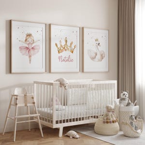 May include: Three framed watercolor prints with a ballerina, a gold crown with the name "Natalie", and a swan. A white crib, high chair, and baskets with stuffed animals complete the nursery decor.