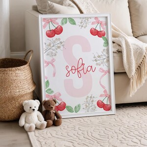May include: A white framed print with a pink letter S and the name "sofia" written in red cursive. The print is surrounded by a border of green leaves, pink bows, and red cherries.
