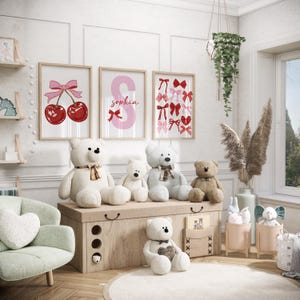 May include: A nursery room with a white, wooden chest with four drawers. There are five teddy bears sitting on top of the chest. Three of the bears are white, one is brown, and one is gray. There are three framed prints on the wall behind the chest. The prints feature a red cherry with a pink bow, a pink letter S with the name "Sophia" written on it, and a pink and red bow pattern.