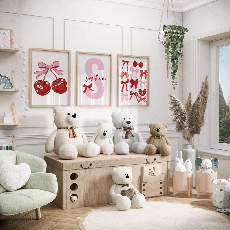 May include: A nursery room with a white, wooden chest with four drawers. There are five teddy bears sitting on top of the chest. Three of the bears are white, one is brown, and one is gray. There are three framed prints on the wall behind the chest. The prints feature a red cherry with a pink bow, a pink letter S with the name "Sophia" written on it, and a pink and red bow pattern.