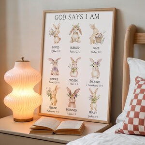 May include: A framed print with the text "GOD SAYS I AM" and illustrations of bunnies with floral accents. The print includes words like "LOVED," "BLESSED," and "SAFE," with corresponding Bible verses. A lamp and open book are in the foreground.