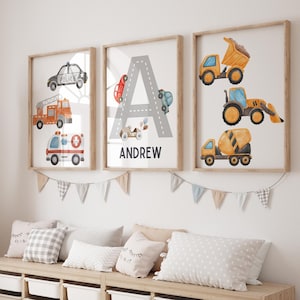 May include: Three framed watercolour prints featuring a letter A with various vehicles, a fire engine, a police car, an ambulance, a cement mixer, a bulldozer, and a dump truck. The middle print has the name Andrew below the letter A.
