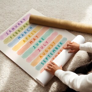 May include: A colorful calendar print with months of the year, each in a different pastel color. The print is rolled up with a brown cardboard tube. The image shows a person holding the rolled-up print.
