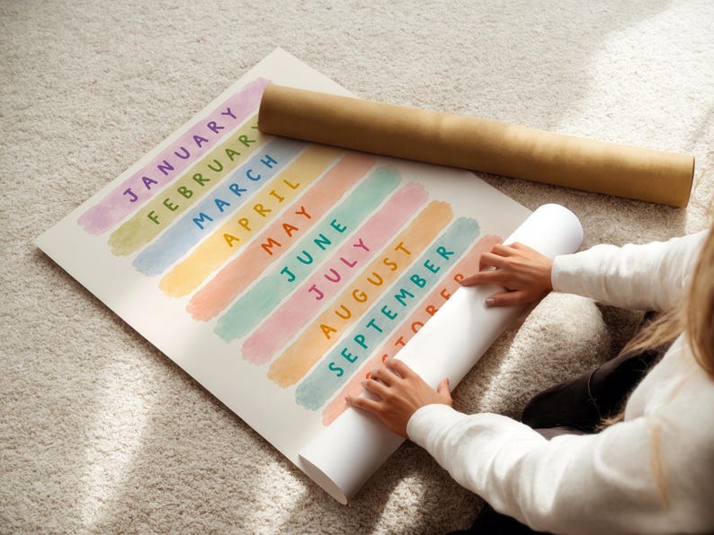 May include: A colorful calendar print with months of the year, each in a different pastel color. The print is rolled up with a brown cardboard tube. The image shows a person holding the rolled-up print.